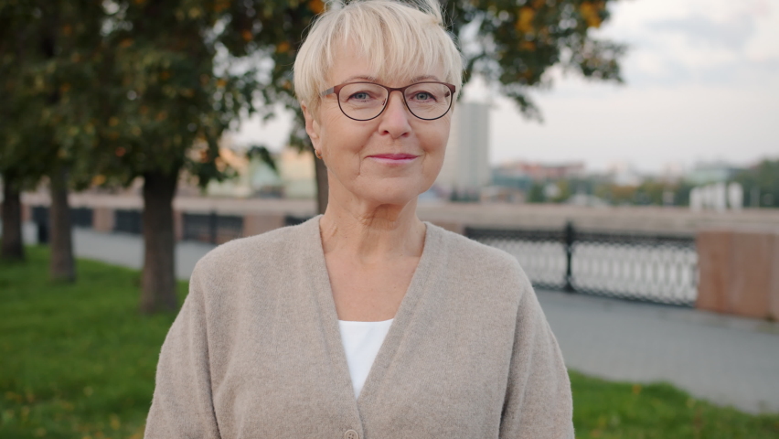 Slow motion portrait of elegant elderly woman smiling looking at camera standing outdoors in urban street. Senior people and emotions concept.