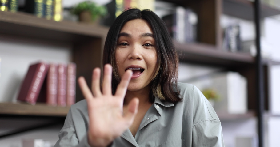 Cute and young Asian woman with braces joining video call session. Beauty girl raises hand greeting and begins talking to friend chat online dating with happy face. Millennial lifestyle concept.