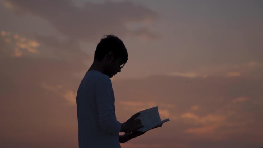 Silhouette of a young Indian student wearing glasses reading a book. School going kid holds book in his hand and reads it. Education concept. Smart Kid is studying hard to improve his future. | Shutterstock HD Video #1079334662