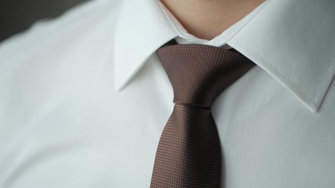 A man in a white shirt ties a brown tie around his neck. learning tying a necktie