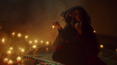 Witch woman cutting finger with knife for sacrifices, sitting in pentagram circle. Sorceress making rite at night, using blood and black witchcraft, cow skull, candles on floor. Halloween concept.
