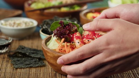 Putting cooked poke bowl on table close-up. Traditional Hawaiian dishes served with sliced vegetables, fish and greenery, dried seaweed. Healthy vegetarian food. Asian vegan raw meal, chopsticks.