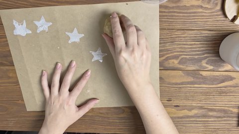 Woman dips potato stamp in form of star in acrylic paint to do print on craft paper. Girl hands decorate wrapping paper on wooden table. Diy and handmade on quarantine.