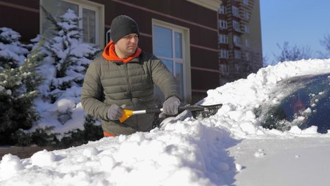A guy cleans fresh snow from his car with a brush on a bright sunny day.Cleaning the car roof and mirror in winter weather from snow and ice. The streets are covered with snow and snowdrifts.