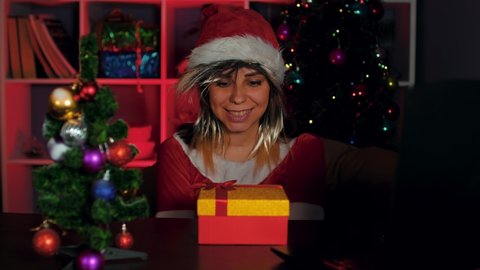 A young woman in a Santa Claus costume in the office opens a gift containing money. A smiling manager in a New Year's suit and hat, holding a stack of money
