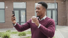Side view of smiling young African American man having video chat on phone outside business office, waving hand, holding coffee. Having stylish clothes. Lifestyle, communication concept. 