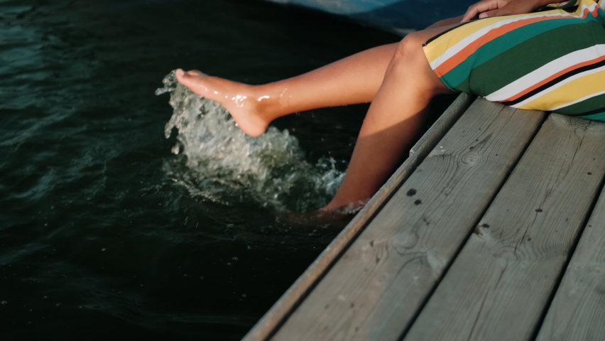 Beautiful African-American girl with dreadlocks in a bright dress sits on a pier among the boats with her legs lowered into the water.
