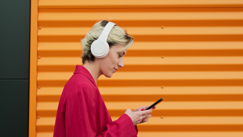 Stylish young woman walking along city street against gray and orange wall using smart phone. Female listening to music with headphones outdoors. Tracking shot. Close-up in 4K, UHD