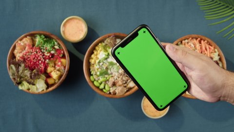 Hawaiian poke bowls, holding smartphone with chroma green screen top view. Cooked poke made of sliced vegetables, seafood and greenery. Healthy vegetarian dishes. Asian vegan raw meal, chopsticks.