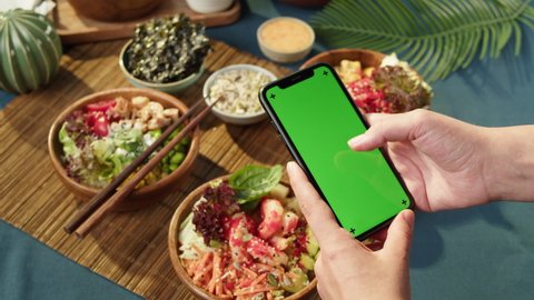 Hawaiian poke bowls, holding smartphone with chroma green screen close-up. Cooked poke made of sliced vegetables, seafood and greenery. Healthy vegetarian dishes. Asian vegan raw meal, chopsticks.