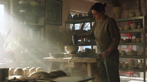 Woman potter is working in pottery workshop, drying raw clay pot with an electric dryer before putting it to a kiln, ceramist lifestyle, beautiful pottery studio, Slow motion.
