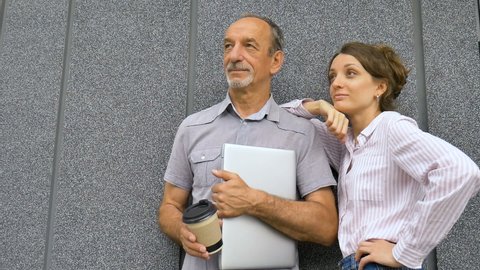 Young woman and senior man are waiting for a meeting standing near the grey wall outdoors in the morning. Colaboration between older coworker and younger colleage