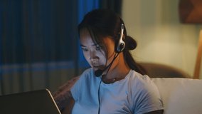 Asian woman in headset smiling and speaking while making video call via laptop near child in dark living room at night at home