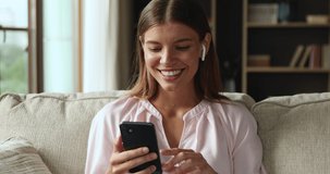 Video call. Laughing female teenager in wireless headset rest on sofa at home speak by phone wave hand say hello to chat buddy. Positive young woman talk to friend on virtual meeting in app having fun