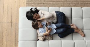 Happy young asian vietnamese mother lying on cozy sofa with small adopted child son, using smartphone software applications together, playing mobile games, watching funny videos online, top view.