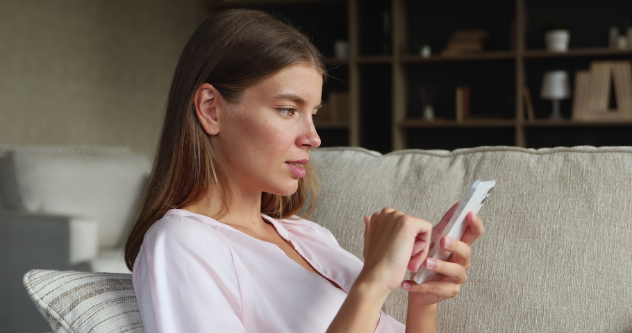 Side view of gen z teenage female satisfied user of modern mobile phone reclining on couch browsing internet downloading software. Young lady surfing social networks on cell choose goods at web store Royalty-Free Stock Footage #1079380448