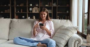 Smiling generation z woman relax on soft couch using smartphone surf social media web pages read news online chat in mobile dating app. Calm young casual lady enjoy networking in virtual space on cell