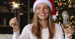 Merry Christmas. Close up portrait of happy young lady hold sparkler champagne record video blog near Xmas tree. Joyful teen female in Santa hat look at camera greet friends with New Year by videocall