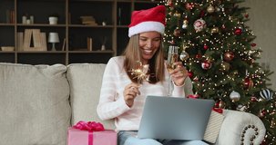 Virtual party. Overjoyed gen z woman in Santa cap celebrate New Year at home by laptop hold sparkler drink champagne talk in conference app. Laughing teen female wish friends a merry Christmas online