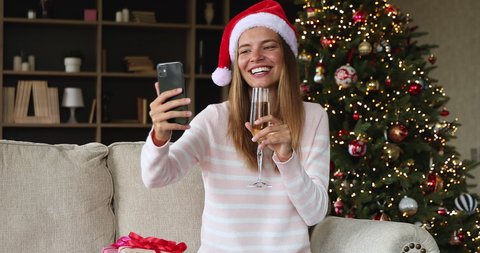 Social distancing and New Year. Happy young lady sit on sofa hold phone make record of virtual Xmas speech. Merry teen female in Christmas hat raise up glass of champagne give best wishes by videocall