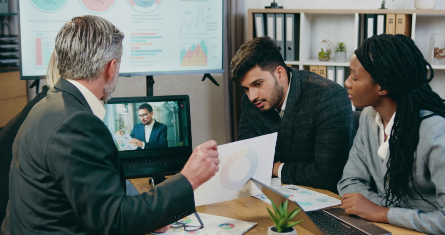 Attractive confident skilled diverse business team explaining report with chart to their bearded head of company during online video chat on laptop in office room