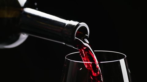 Red wine pouring from bottle into goblet. Close-up of red wine pouring in wine glass at black background. Slow motion. Macro shot