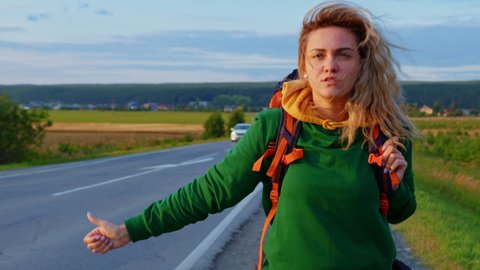 Beautiful blond girl with backpack hitching ride. Young woman hitchhiker steps back slowly along roadside, waits for passing car to stop. Hitchhiking in countryside. Hiker, tourist, traveller