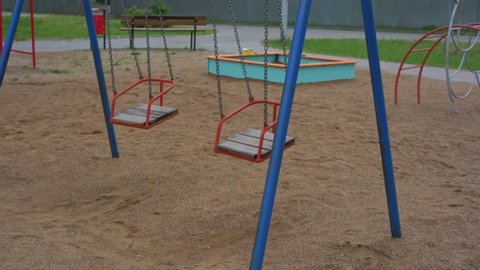 Wind sways red metal wooden swings on chains on empty children playground with sandpit ladder and bench in morning slow motion