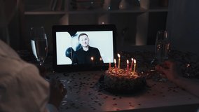 Birthday video call. Virtual celebration. Remote holiday meeting. Female friends with champagne watching man greeting speech on tablet screen on decorated table with cake.