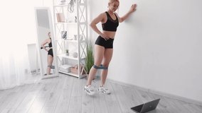 Enjoying fitness. Athletic woman. Home sport. Online lesson. Happy sportive lady doing legs aside training with rubber bend workout laptop on floor light room interior copy space.