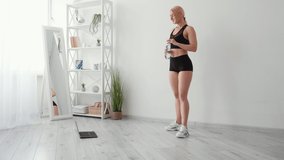 Hydration body. Sportive woman. Online training. Home gym. Happy athletic lady in black sportswear drinking water before doing warming exercise laptop on floor light room interior.