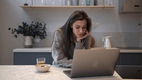 Brunette woman working in kitchen, drinking glass of a hot coffee and plate on the table. Talking on a video call in laptop at modern room. Concept of breakfast in the morning, remote work