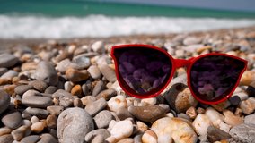 Sunglasses in the form of hearts lies on a pebble beach with sea waves in the background. The concept of a great vacation. 4k video.