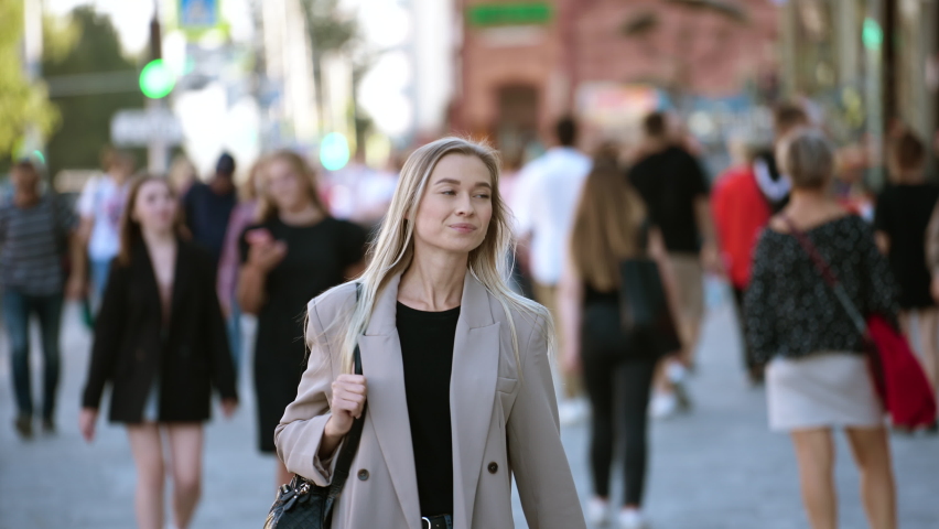 Attractive natural haired blonde colored European looking woman walking in streets of modern downtown with smiling, smirking face. Stylish trendy businesswoman having a walk on crowded city streets.