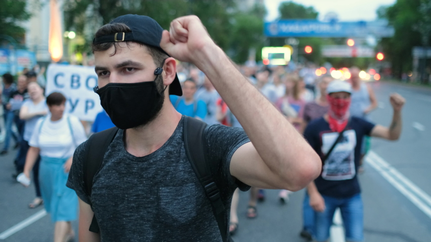Masked face of male protester rebel in coronavirus restriction resistance crowd picket demonstration. Political opposition activist guy in covid-19 facemask waves arm fist. City riot, rally revolt. Royalty-Free Stock Footage #1079407232