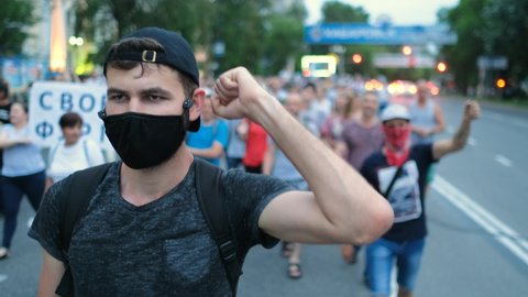 Masked face of male protester rebel in coronavirus restriction resistance crowd picket demonstration. Political opposition activist guy in covid-19 facemask waves arm fist. City riot, rally revolt.