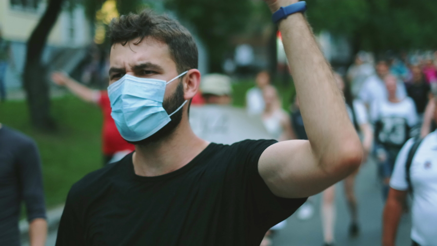 Masked face of male protester rebel in coronavirus restriction resistance crowd picket demonstration. Political opposition activist guy in covid-19 facemask waves arm fist. City riot, rally revolt. Royalty-Free Stock Footage #1079407244