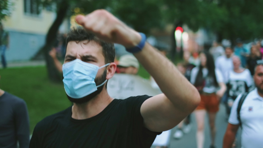 Masked face of male protester rebel in coronavirus restriction resistance crowd picket demonstration. Political opposition activist guy in covid-19 facemask waves arm fist. City riot, rally revolt. Royalty-Free Stock Footage #1079407244