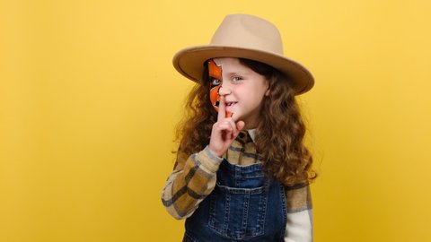 Secret charming fun curly little girl with Halloween makeup mask wears brown hat, say hush be quiet with finger on lips shhh gesture, posing isolated on yellow studio background with copy space 