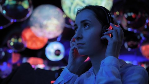 Woman wearing wireless black headphones and looking around in dark room of interactive museum or exhibition with colorful illumination. Immersive, futuristic, entertainment concept