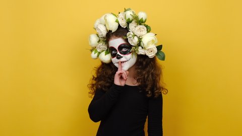 Secret little girl child with creative Halloween or day of death makeup mask wears flowers wreath, say hush be quiet with finger on lips shhh gesture, posing isolated over yellow studio background