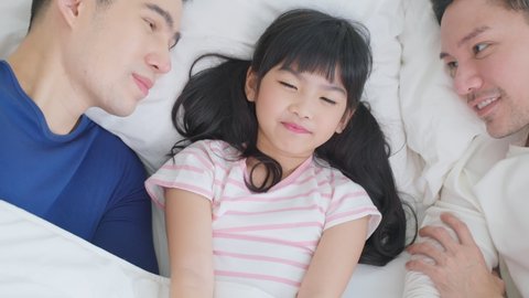 Portrait of handsome man gay family with young kid daughter in bedroom. Attractive romantic male lgbt couple lying down on bed and kiss little adorable girl child in morning, enjoy parenting in house.