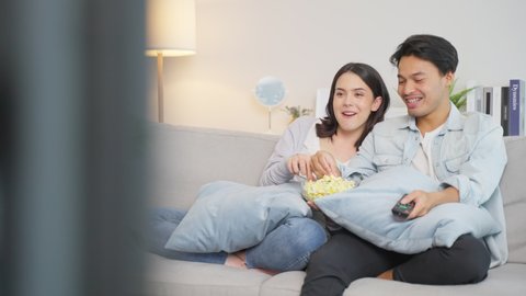Asian young loving couple watch movie together in living room at home. Attractive man and woman sit on sofa, Happy boyfriend use remote control TV show and having fun laughing with girlfriend in house