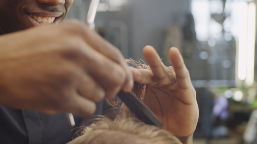 Close up shot of young handsome black barber smiling and talking while cutting hair of client with shears and comb in barbershop | Shutterstock HD Video #1079417672