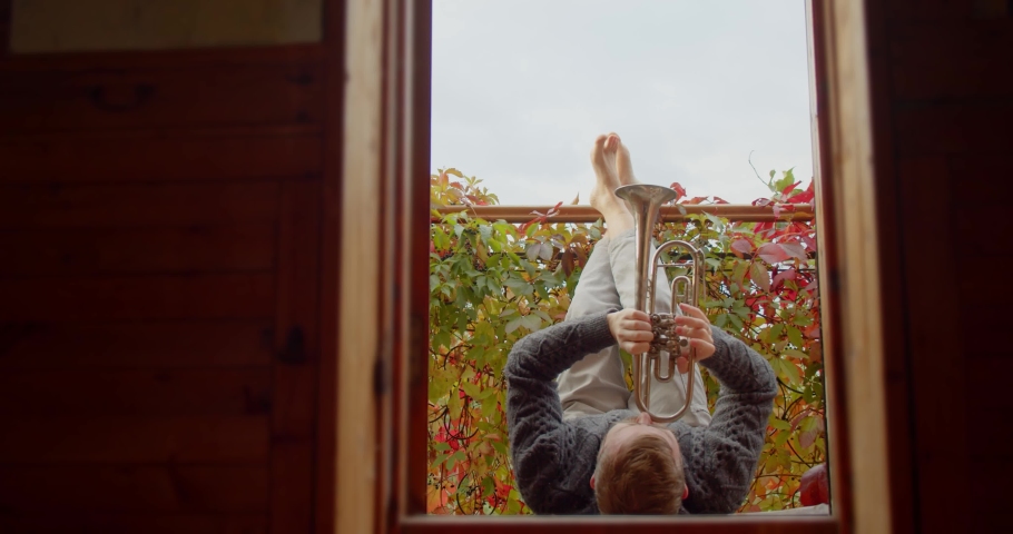 man lies on the balcony and plays the musical trumpet against the background of autumn foliage. 