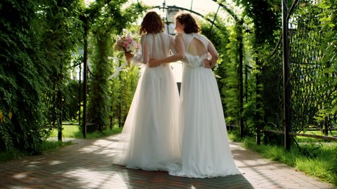TRACKING Candid shot of two happy lesbian LGBT brides wearing boho dresses walking through alley on their wedding day. Shot with 2x anamorphic lens