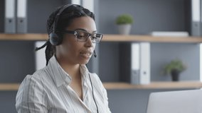 Semi profile portrait of young confident african american woman call center operator wearing headset talking with client via video chat on laptop, slow motion