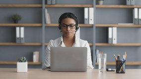 Online business negotiations. Young african american lady sales manager wearing headset talking with client via video call on laptop, sitting at office, slow motion