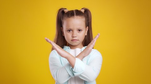 Stop children abusement and bullying. Serious cute little schoolgirl crossing hands and shaking head no, expessing rejection and denial, orange studio background
