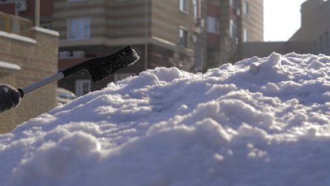 A guy cleans fresh snow from his car with a brush on a bright sunny day.Cleaning the car roof and mirror in winter weather from snow and ice. The streets are covered with snow and snowdrifts.